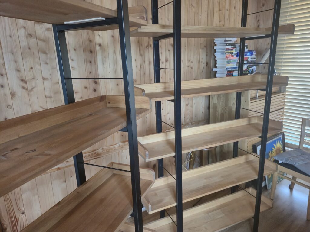 A giant empty bookshelf set combining a black iron frame and sturdy solid wood shelves, purchased from Cheongju Station Furniture Plaza and installed against the wood-paneled wall of the room.